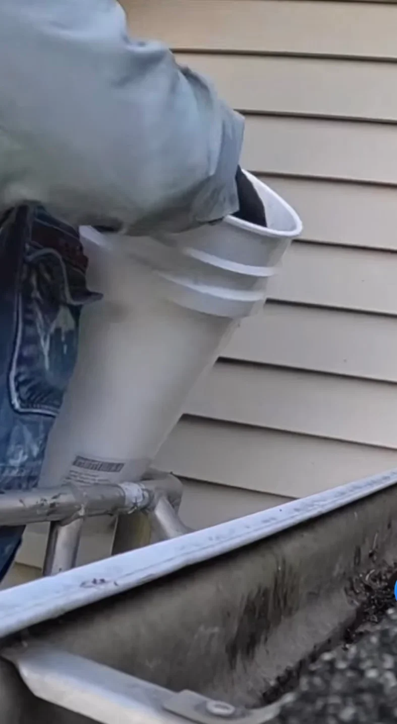 A technician from American Power Washing & Exterior Services clearing packed oak and maple leaves from a residential gutter on a Madison, WI home. Clean downspout visible with water flowing freely after service.